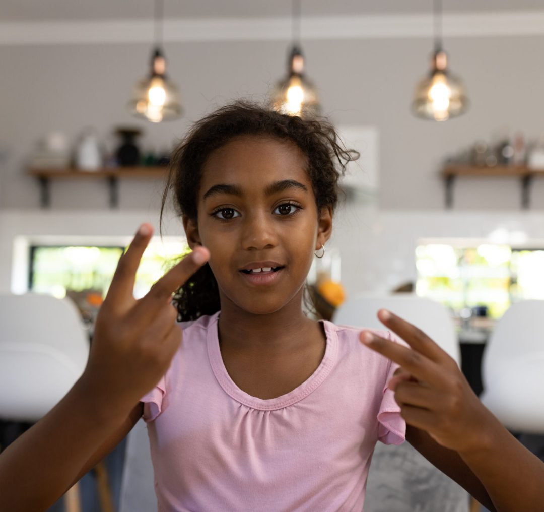 Portrait of happy biracial girl making video call at home, gesturing with fingers and smiling. Childhood, communication, domestic life and technology.